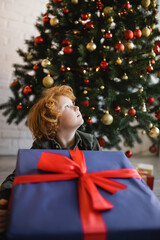 Wall Mural - redhead child looking away near huge gift box and decorated Christmas tree