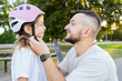 © Julija - Man helps child girl fastens protective helmet for learning to ride skateboard at park. Father helping his daughter to wear a cycling helmet.