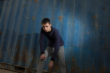 young caucasian guy posing with a serious face dressed in a sweater and jeans in an industrial area against a blue iron wall and a blue cloudy sky. lower angle