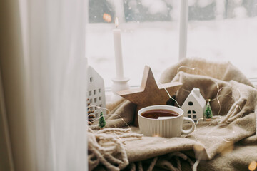 Wall Mural - Cozy home at snowy day. Warm cup of tea, candles, lights, little christmas houses and trees, wooden star on cozy blanket on windowsill. Winter hygge. Atmospheric scandinavian mood