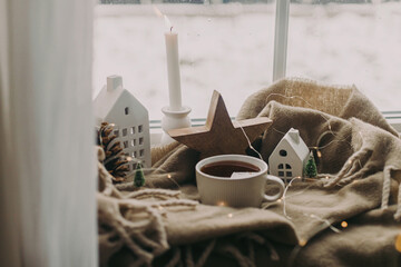 Wall Mural - Winter hygge. Warm cup of tea, candles, lights, little christmas houses and trees, wooden star on cozy blanket on windowsill. Cozy home. Atmospheric snowy day. Christmas background