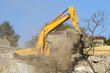 Yellow crawler excavator demolishes dilapidated real estate for future construction of new modern house. Clouds of dust around special construction equipment cleaning up rubble of collapsed building