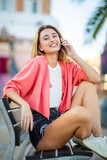 Fototapeta Przestrzenne - Smiling woman in stylish summer clothes talking on the phone sitting on a bench