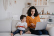 © Iona - Cheerful young Italian woman with curly hair dressed in orange blouse and blue pants sitting on sofa using laptop, working at home at the kitchen with son playing on phone, talking with mom. Family
