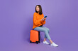Full length photo of pretty young woman sit suitcase hold device dressed stylish orange knitted outfit isolated on purple color background