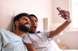 Young Couple At Home Posing For Selfie On Mobile Phones In Bed Together