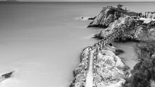 Grayscale Shot Of A Footbridge Along Big Rocks On A Coast