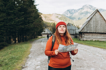 Wall Mural - Stylish woman holding paper map relaxing in nature. Travel and active lifestyle concept. Hiking active female lifestyle wearing backpack and red knit hat exercising outdoors.