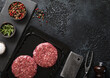 © DenisMArt - Raw beef burgers sealed in vacuum tray with fork and cleaver on wtone board with salt and pepper on black background.