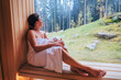 © Soloviova Liudmyla - Relaxing Woman wrapped white towel sitting on the wooden bench in Hot Finnish sauna with a huge wide window with green forest view and enjoying pleasant healthy body care temperature treatment.