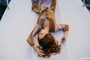 Gorgeous red haired woman laying on yacht with wavy loose hair, looks satisfied and attempting to recharge herself. Attractive Swedish woman on vacation at sea enjoying careless life. Rest and travel