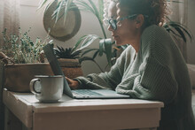 Online Activity At Home With Woman Using Laptop Sitting On The Floor On A Little Table With Green Garden Plants In Background. Internet Connection. Female People Surf The Web Searching Job And Infos