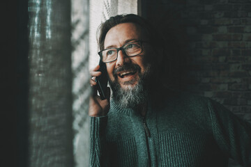 One happy young mature man speaking at the phone smiling and having fun at home near the window. Concept of people and mobile cell communication. Male person using cellular to communicate with friends