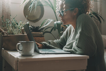 Wall Mural - Online activity at home with woman using laptop sitting on the floor on a little table with green garden plants in background. Internet connection. Female people surf the web searching job and infos
