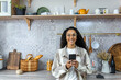 © Liubomir - A beautiful young Latin American woman is holding a phone in her hands. Standing at home in the kitchen, looking at the camera, smiling.