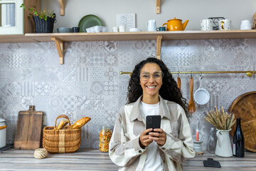 A beautiful young Latin American woman is holding a phone in her hands. Standing at home in the kitchen, looking at the camera, smiling.