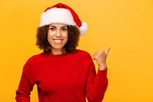 Christmas, Xmas Concept. Happy Smiling Woman In Santa Claus Hat Pointing Finger At Copy Space On Orange Background, Looking At The Camera