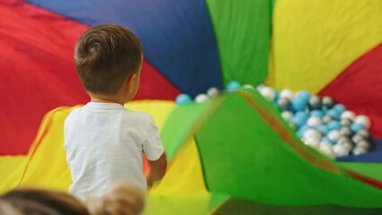 Wall Mural - little Caucasian boy playing with a colorful piece of material and many balls on it, full shot kindergarten. High quality 4k footage