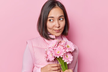 Wall Mural - Portrait of pensive young Asian woman with thoughtful expression wears turtleneck and vest holds bouquet of gerbera flowers celebrates birthday isolated over pink background has romantic mood