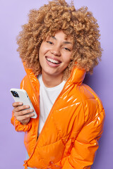 Wall Mural - Happy curly teennage girl dressed in warm orange jacket smiles broadly holds cellphone checks notification going to have walk outdoors expresses positive emotions isolated over purple background