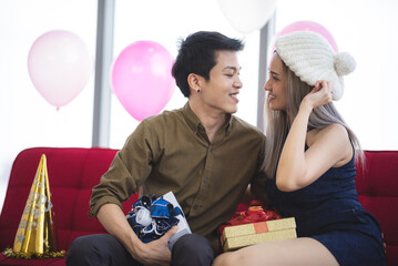 Wall Mural - Handsome young birthday man wearing cap holding and opening gift while sitting besides beautiful girlfriend holding gift and teasing him by pulling his ears and sitting on couch at home party