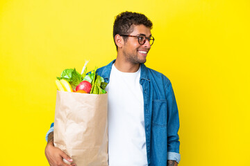 Young Brazilian man holding a grocery shopping bag isolated on yellow background looking to the side and smiling