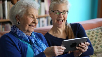 Happy elderly senior woman and mature woman look at tablet computer while drinking tea on a couch in a living room.