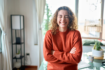 Young smiling pretty lady having fun feeling joyful at home, happy beautiful positive professional woman wearing orange sweater standing in modern cozy apartment living room or in office.
