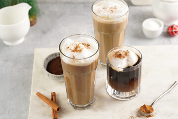 Wall Mural - Three tall glasses with warm coffee drink with cinnamon, whipped milk foam and caramel on a marble board and christmas ornaments and decoration