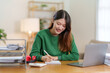 Young Asian business woman sitting happily working on a laptop take notes carefully and smile happily on assignment at home.