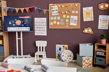 Part of spacious classroom for primary school children with pictures and notes on boards, paintings, chair and boxes standing by wall