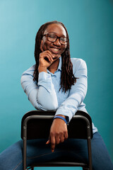 Wall Mural - Excited cheerful african american pretty attractive woman sitting on chair while smiling at camera on blue background. Joyful pleased young adult person with confident smile and casual looking.