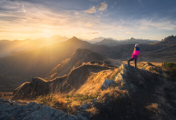 Wall Mural - Young woman on mountain peak and beautiful mountains in haze at colorful sunset in autumn. Dolomites, Italy. Sporty girl, mountain ridges in fog, orange grass, trees, golden sun in fall. Hiking	