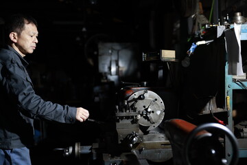 A craftsman poses in gray work clothes in front of a lathe at a local factory. Conceptual images of the essence of manufacturing, technical succession, and the challenge of high-precision machining.