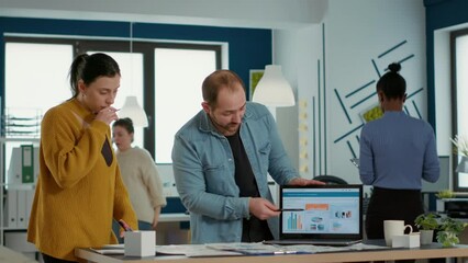 Wall Mural - Startup office worker explaining sales startegy to new employee using laptop with business analytics and turnover charts. Man standing at desk talking about marketing with woman doing internship.