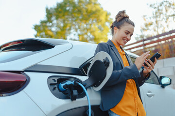 young woman with smartphone waiting while her electric car charging in home charging station, sustai