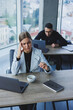 © Дмитрий Ткачук - A female manager sits at a desk using a laptop in a modern office with a colleague in the background. Working atmosphere in an office with large windows.