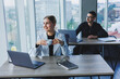 © Дмитрий Ткачук - A female manager sits at a desk using a laptop in a modern office with a colleague in the background. Working atmosphere in an office with large windows.