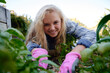 © StratfordProductions - Happy young caucasian woman in checked shirt and gardening gloves by plants in plant nursery