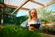© StratfordProductions - Young caucasian woman in checked shirt using digital tablet by plants at plant nursery