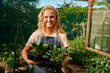 © StratfordProductions - Happy young caucasian woman in apron looking at camera and holding plants at plant nursery