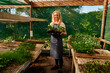 © StratfordProductions - Young caucasian woman wearing apron looking at camera and holding plants at plant nursery