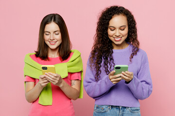 Young two friends smiling happy fun women 20s wears green purple shirts together hold in hand mobile cell phone use internet chat isolated on pastel plain light pink color background studio portrait.