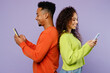 © ViDi Studio - Young couple two friends family man woman of African American ethnicity wear casual clothes together hold in hand use mobile cell phone stand back to back isolated on pastel plain purple background.
