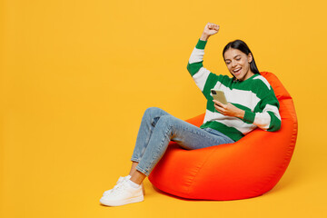 Full body young latin woman wear casual green knitted sweater sit in bag chair hold in hand use mobile cell phone do winner gesture isolated on plain yellow background studio People lifestyle concept.