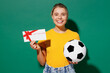 Young happy woman fan wear basic yellow t-shirt cheer up support football sport team hold soccer ball store gift certificate coupon voucher card watch tv live stream isolated on dark green background.