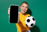 Young woman fan in basic yellow t-shirt cheer up support football sport team hold soccer ball watch tv live stream use mobile cell phone closeup blank screen isolated on dark green background studio