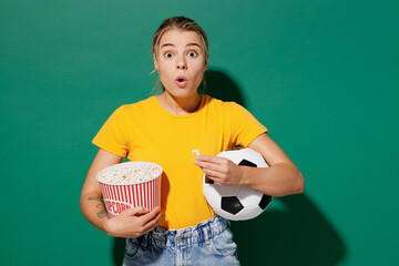 Young amazed woman fan in basic yellow t-shirt cheer up support football sport team eat popcorn hold in hand takeaway bucket soccer ball watch tv live stream isolated on dark green background studio