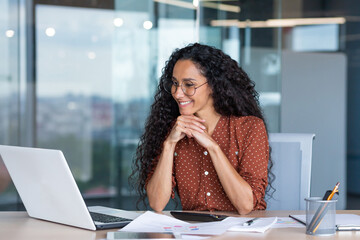 Wall Mural - Young happy and successful businesswoman in glasses working with documents inside office, Hispanic woman with laptop looking at bills and contracts, financier with curly hair using laptop.