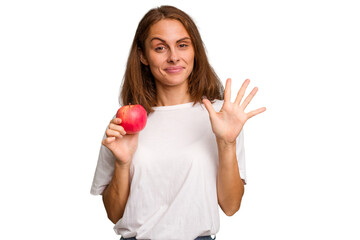 Young caucasian woman holding a red apple isolated smiling cheerful showing number five with fingers.